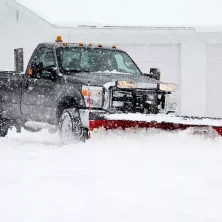 A black truck with a red snowplow from a 'dumpsters rental' service clears snow, parked near a white building.